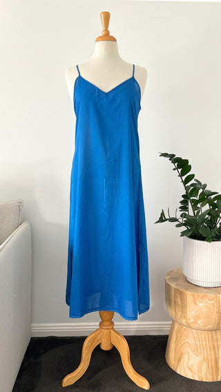 Blue slip dress on a mannequin against a white wall with a plant and wooden stool in the background.